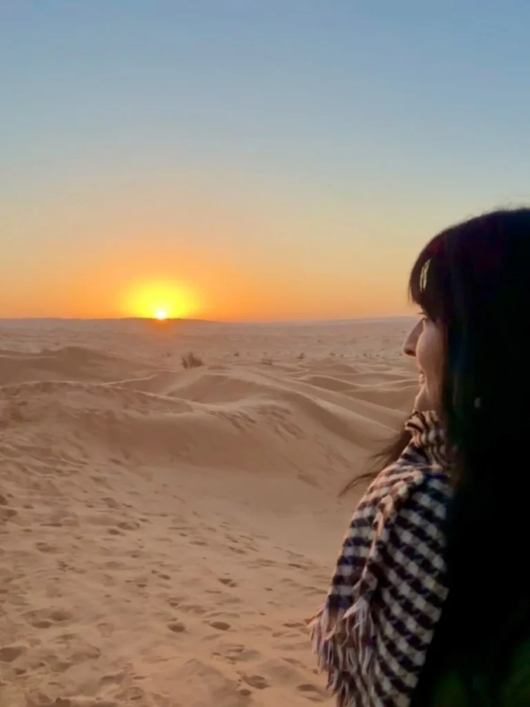 Bejal standing looking at the sunrise at camp Abdelmoula. Bejal is wearing a checked black and white scarf