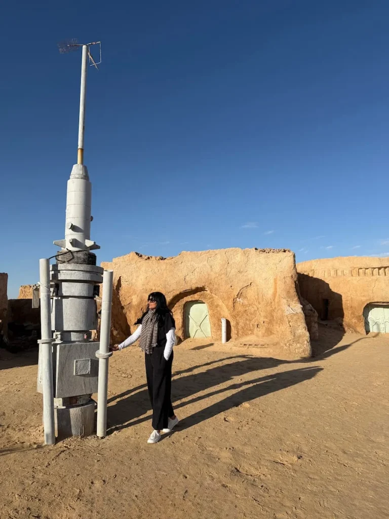 Bejal standing next to one of the spaceships at Mos Espa in Tunisia. There are desert syle small houses/caves in teh background in the sand and the skies are bright blue. Bejal is wearing a black jumpsuit with a checked scarf and white long sleeve t-shirt