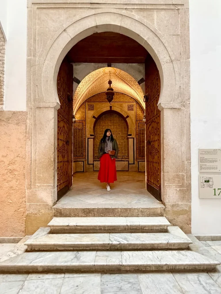 Bejal wearing a red skirt and khaki coat, at old doorway in the Bardo museum, Tunisia