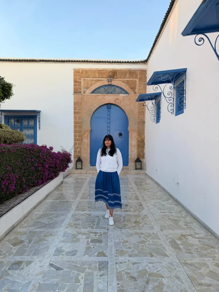 Bejal infront of blue door or Dar Said wearing a blue skirt adn white jumper walking down the hotel walkway, Tunisia