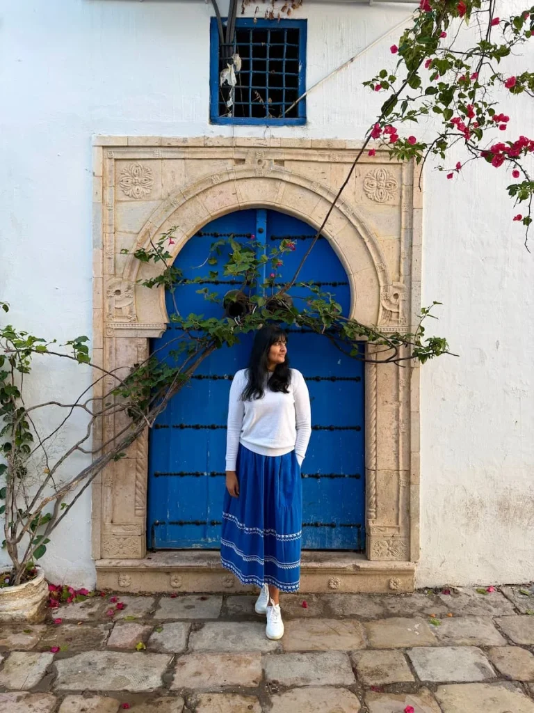 Bejal standing infront of a blue studded door with foliage framing the image. Bejal is wearing a blue skirt and white jumper as well as white trainers. Sidi Bou Said, Tunisia