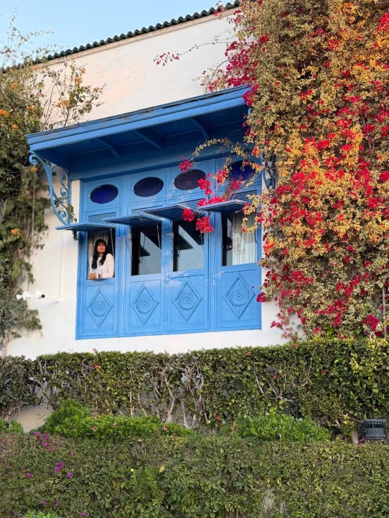 Bejal looking out of the window at Dar Said, Tunisia. The window is bright blue and Bejal is peeking through wearing a white top. the window is surrounded by foliage and bougainvillea