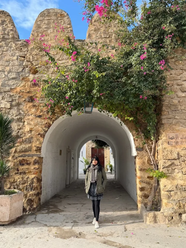 Bejal wearing a green coat and black leggings, standing in the entrance to the stone walled Hammamet Medina