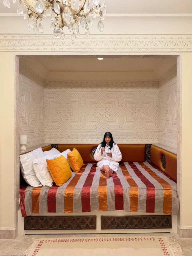 Bejal sitting in bed wearing a fluffy white robe at Dar Alouini, Kairouan. Tunisia. bejal is drinking a cup of tea and the bed has a red and orange stripe bed spread and white and orange linen covered pillows.