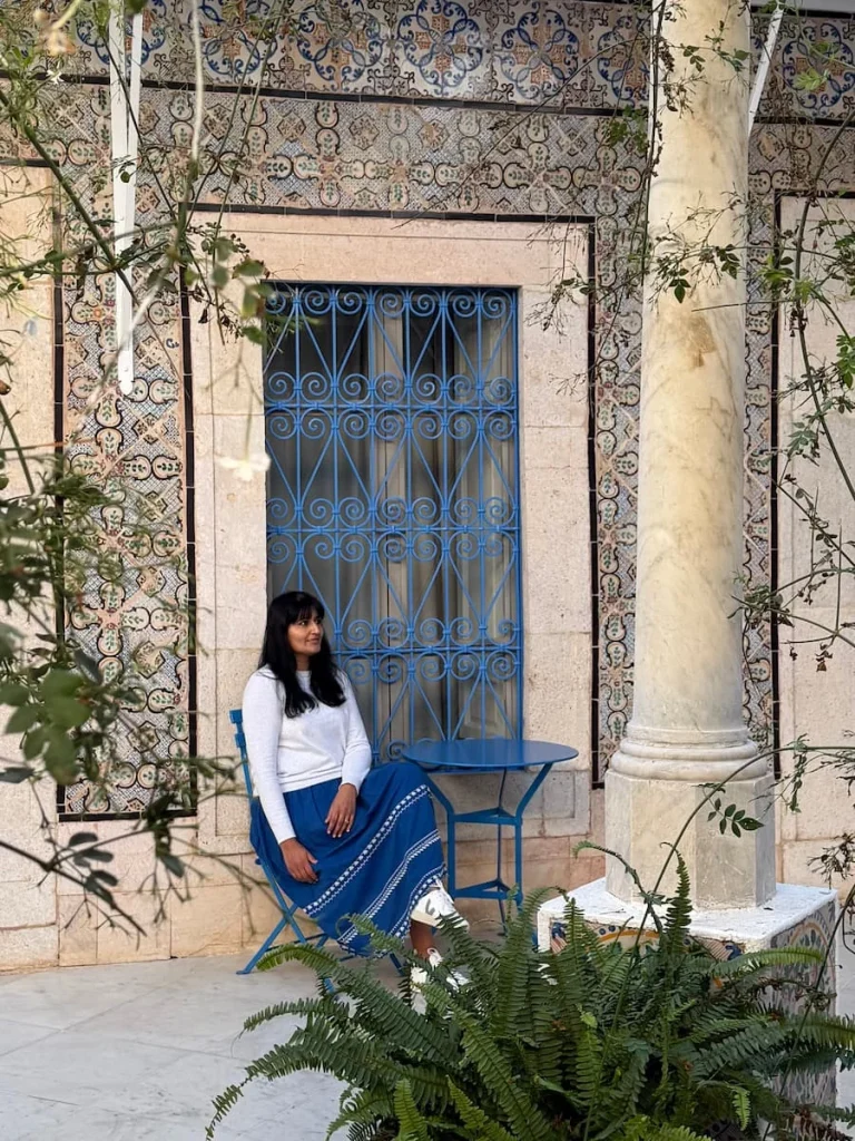 Bejal sitting in the courtyard of Dar Said Hotel, Tunisia wearing a white top and blue skirt with foliage all around