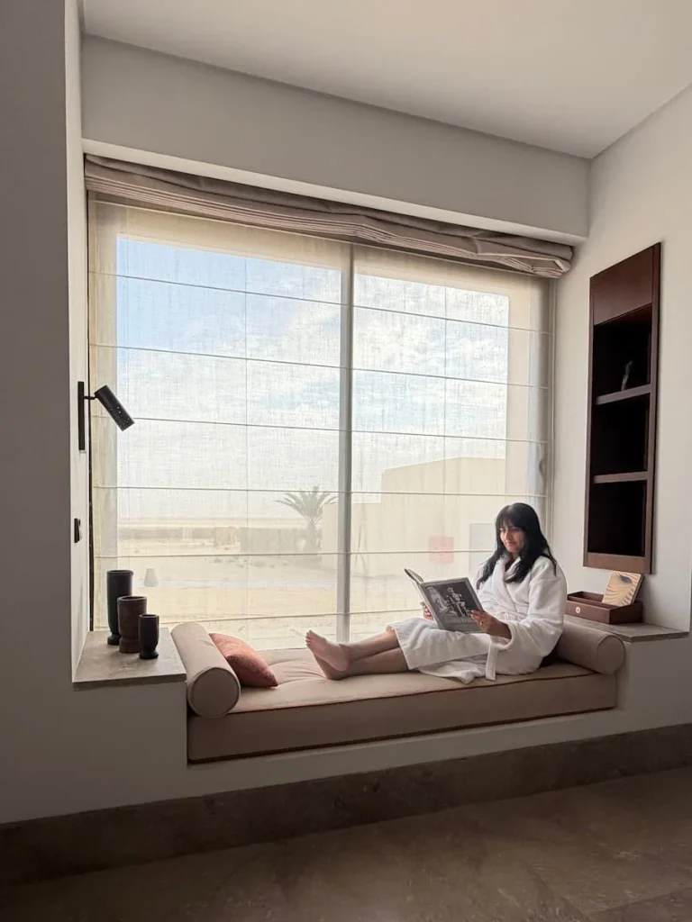 Bejal sitting in the window of the villa at The Mora Sahara Tozeur. The window seat has views out to the desert and has comfy circular cushions at either end. Bejal is wearing a white fluffy robe