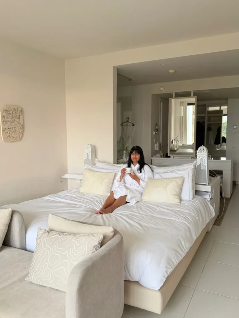 Bejal wearing a white dressing robe sitting in bed at La Badira drinking coffee. The bed has white linens as does the sofa. In the background there is a mirror. La Badira is located in Hammamet