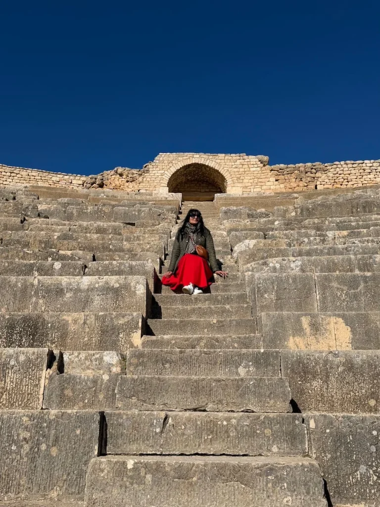 Bejal sitting on steps of amphitheatre wearing a red skirt and jacket, in Dougga, Tunisia. There is a stone archway behind her and bright blue skies.