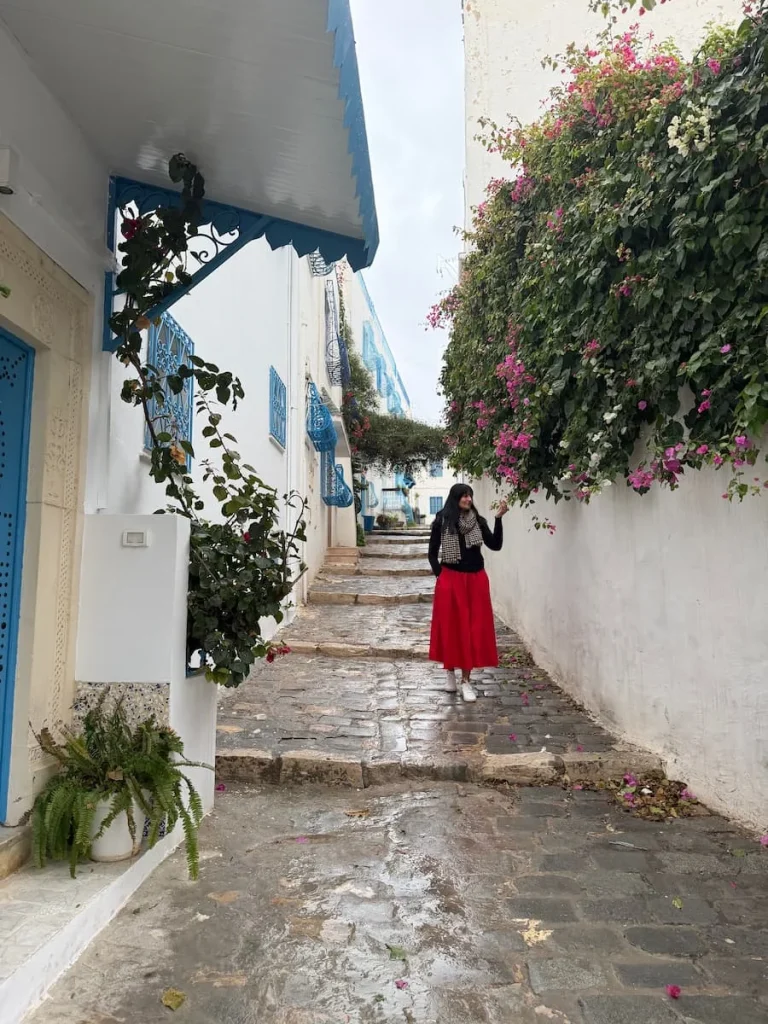 Bejal walking down a white washed alleyway wearing a red skirt with a khaki jacket and smelling teh draping bougainvillaea plants surrounded her in Sidi Bu Said, Tunisia