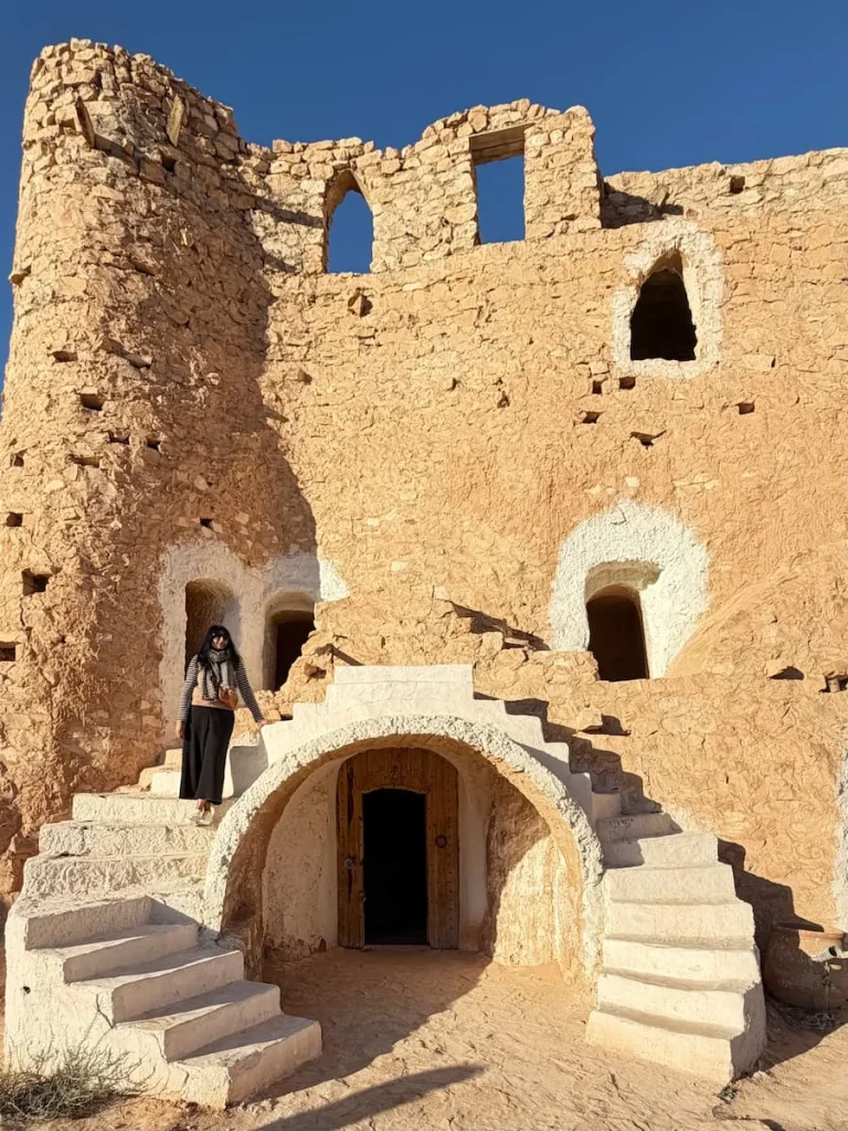 Bejal standing at Troglodyte houses in Matmata, Tunisia. jpeg. These particular houses have been restored to make them look more attractive with white window and door framing. Bejal is standing on the stairs wearing black trousers and a stripey black and white breton