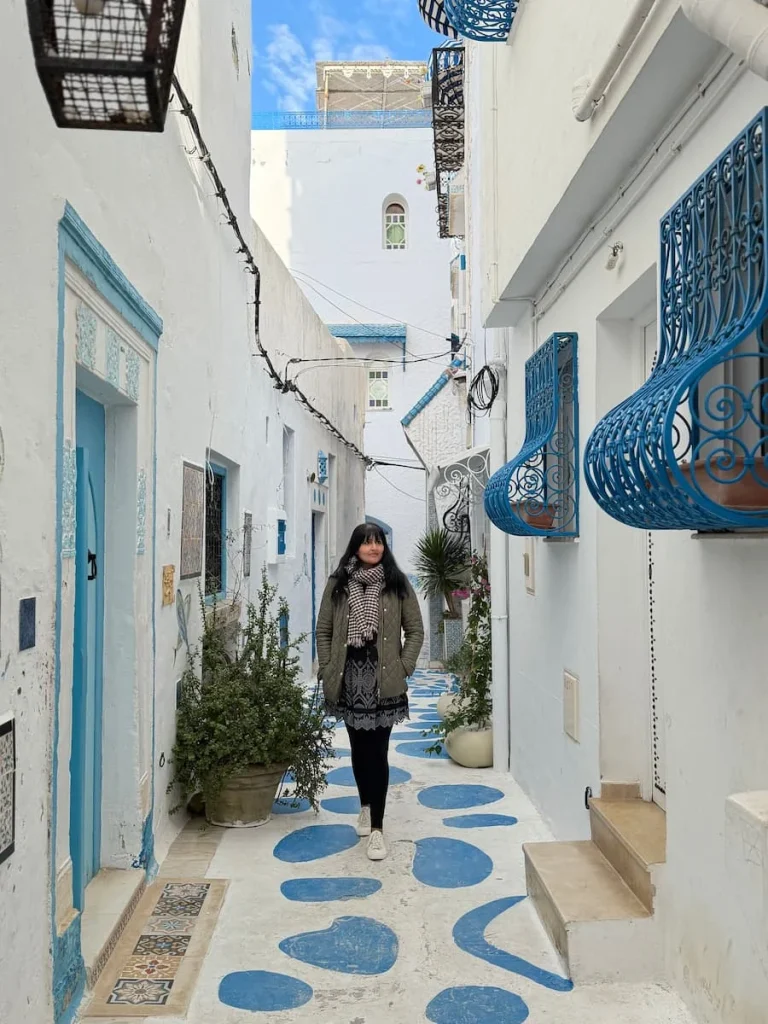 Bejal walking down blue and white alley in Hammamet. Tunisia. Bejal is wearing a khaki jacket with a black short dress and leggings