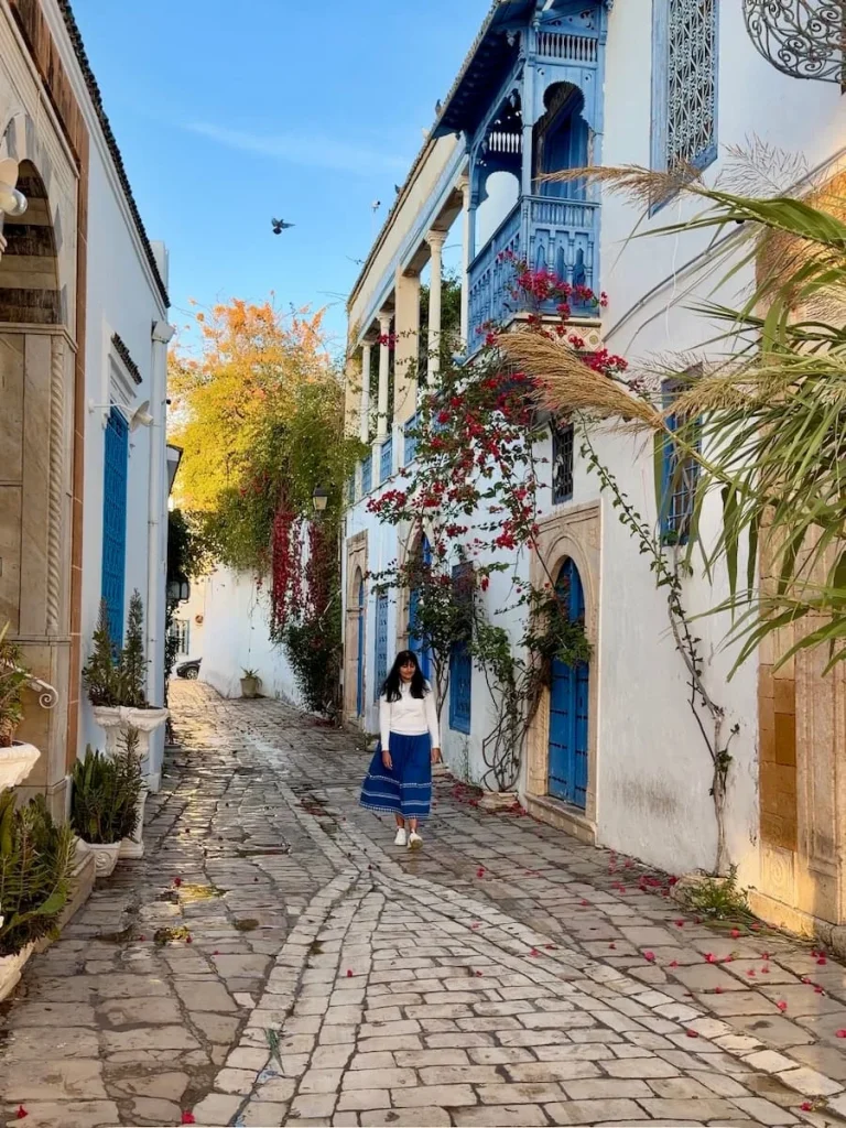 Bejal walking down streets of Sidi Bou Said, Tunisia. Theer are blue doors and bourgainvilia in the background. bejal is wearing a blue skirt and white top
