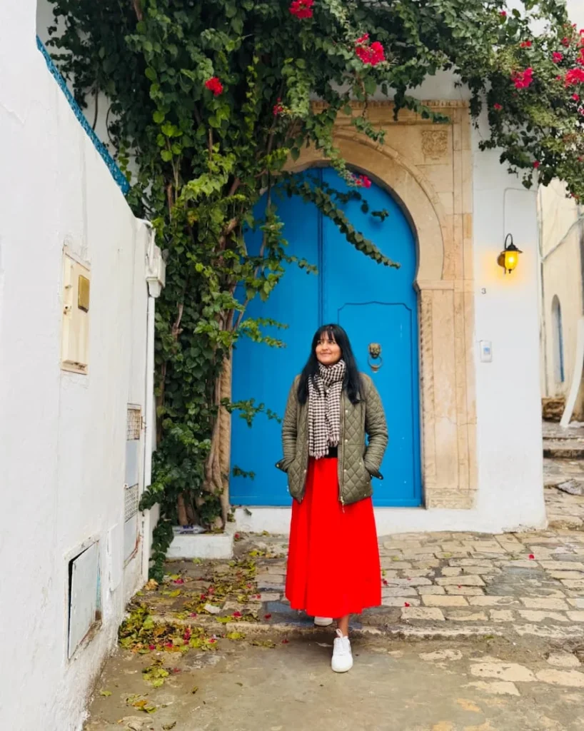 Bejal with red skirt adn khaki jacket infront or blue door in . She is surrounded with bougainvillaea in Sidi Bou Said, Tunisia