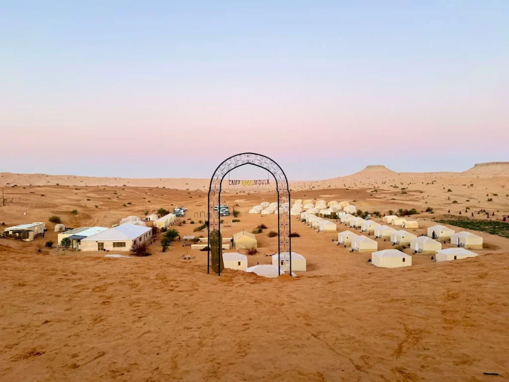 A panoramic image of Camp Abdelmoula in Tembaine with tents and teh desery in the background. Its sunrise and teh site can be seen through the wrought iron archway that says 'Camp Abdelmoula'.