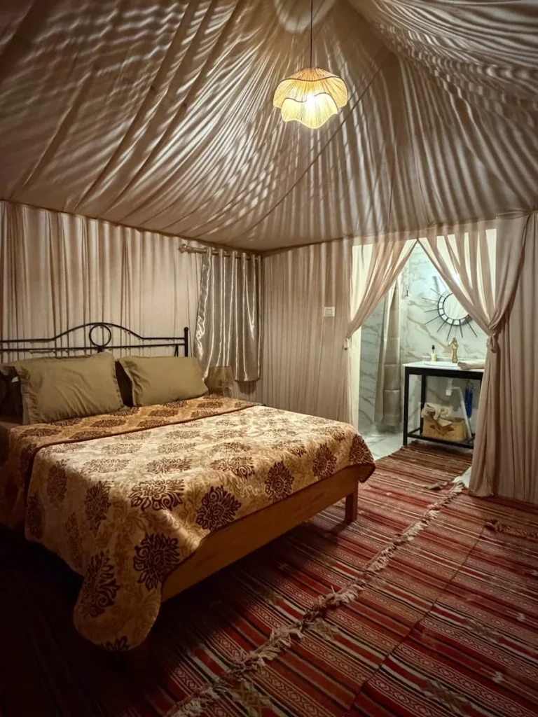The inside of an ensuite tent, with a wrought iron bed, blankets, drapes and a bathroom with a basin, shower and toilet.