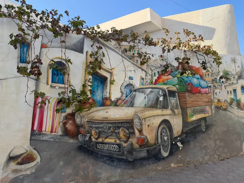 Things to do in Djerba: Djerbahood street art, Djerba Tunisia. The mural is of a colourful truck willed with thing sin the back and is painted against the blue and whitewashed houses.