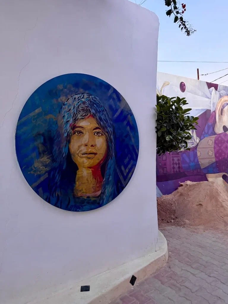 Circularr face street art in Djerbahood of a local lady wearing a blue head dress on a white washed background. 