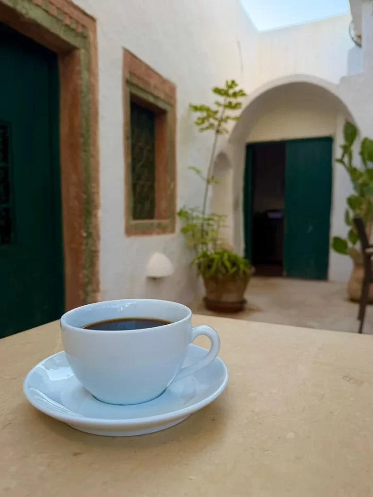 a white coffee cup filled with black coffee at Dar Dhiafa. There is a green door in the background with a trailing pot plant, Djerba
