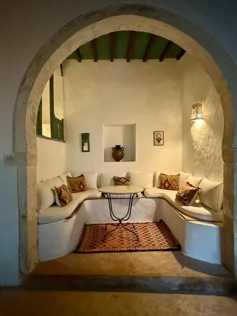 The seating area of a suite in Dar Dhiafa with white washed walls in an alcove with cushioned seats and colourful scatter cushions and low ligting