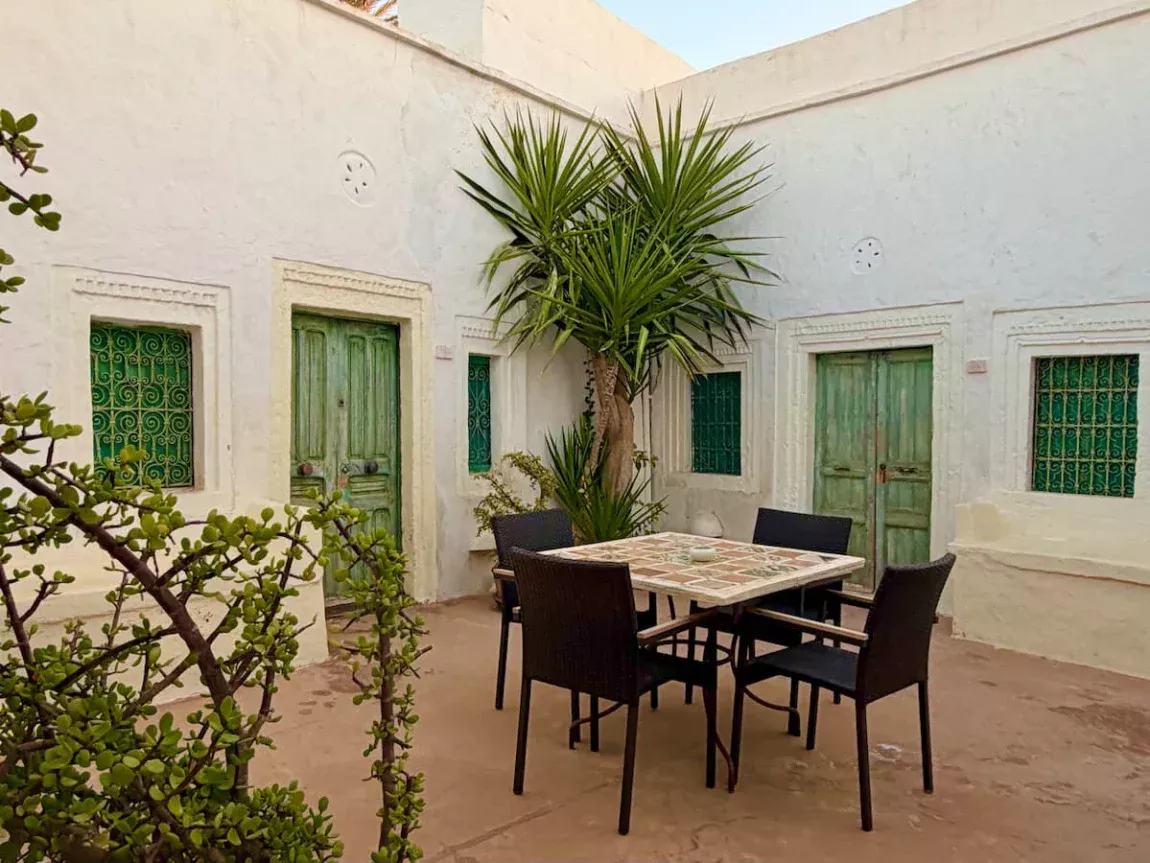 Dar Dhiafa courtyard, Djerba jpeg. There are green suite doors in the background surrounding a table in the centre with chairs. The courtyard is open to the elements and there are potted plants in the corners.