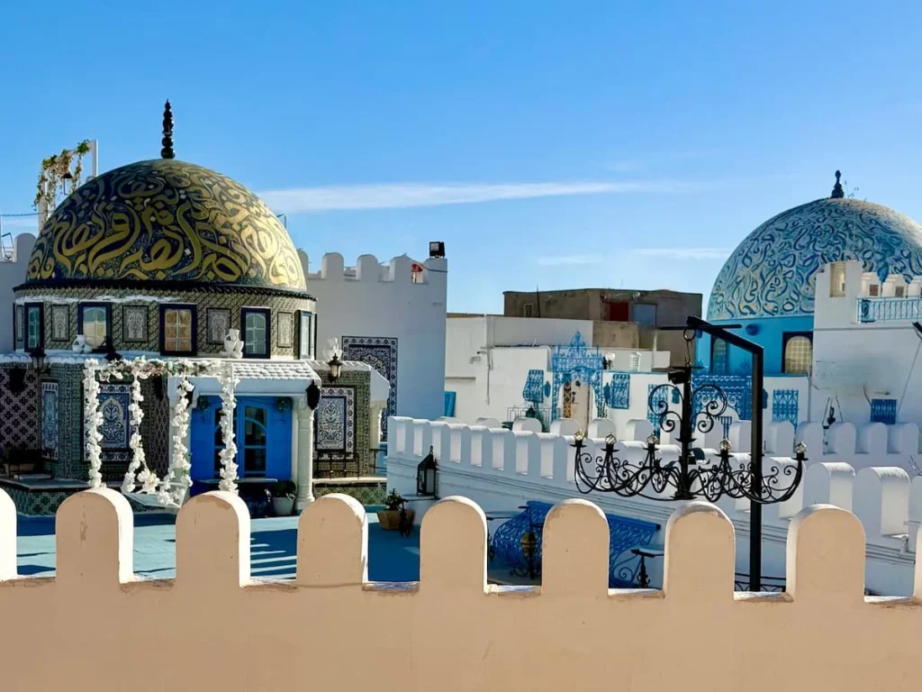 Domed roofs in Kairouan, Tunisia with. gold and blue inscriptions on the domes. There are lights adn a white wall surrounding the rooftops as well as white flowers used as decoration. The sky is bright blue behind the domes.