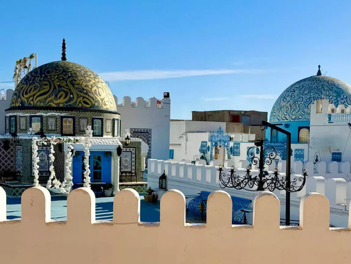 Domed roofs in Kairouan, Tunisia with. gold and blue inscriptions on the domes. There are lights adn a white wall surrounding the rooftops as well as white flowers used as decoration. The sky is bright blue behind the domes.