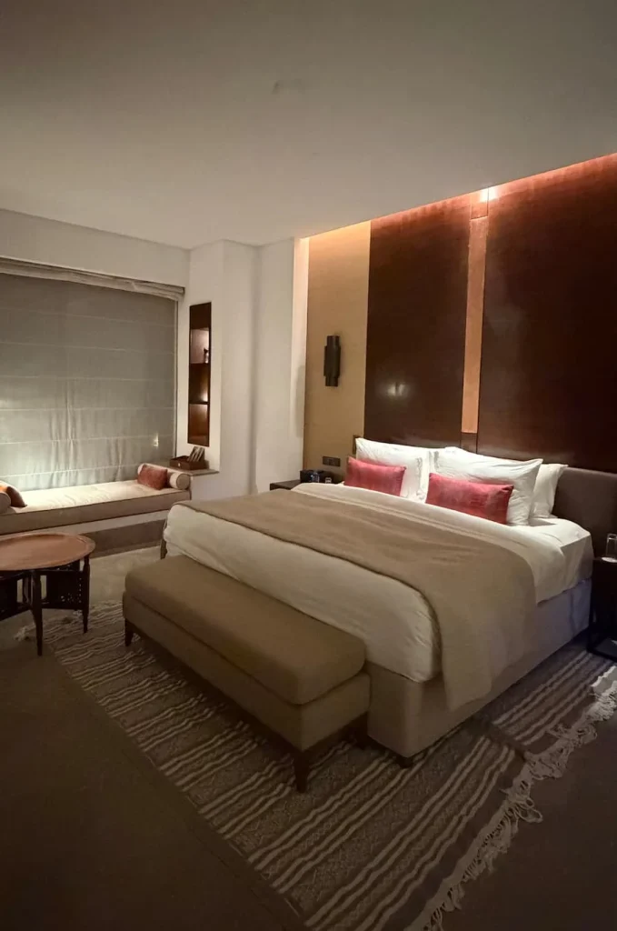 Double bed in the villa at The Mora, Tozeur. The room is lit by discreet atmospheric lighting and tge bed has white linen with red scatter cushions and dark wood side tables