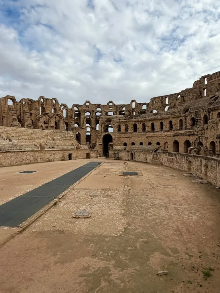 The interiors view of the El Djem Amphitheatre with the left hand side with varying levels of seating. The right has the colosseum structure in tact. There is a blue- grey sky in the background.