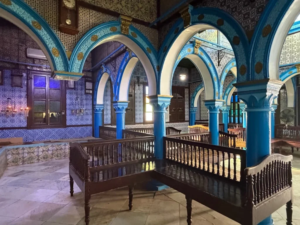 El Ghriba Synagogue interiors that are blue and white arches with grown seating. There is Hebrew writing on the walls