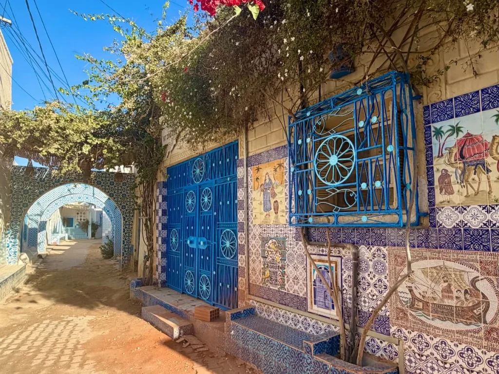 A beautiful Erriadh restaurant, Djerba. It has blue grilled windows and entrance door. The top is draped in foliage and flowers and there is an archway to the right of teh restaurant