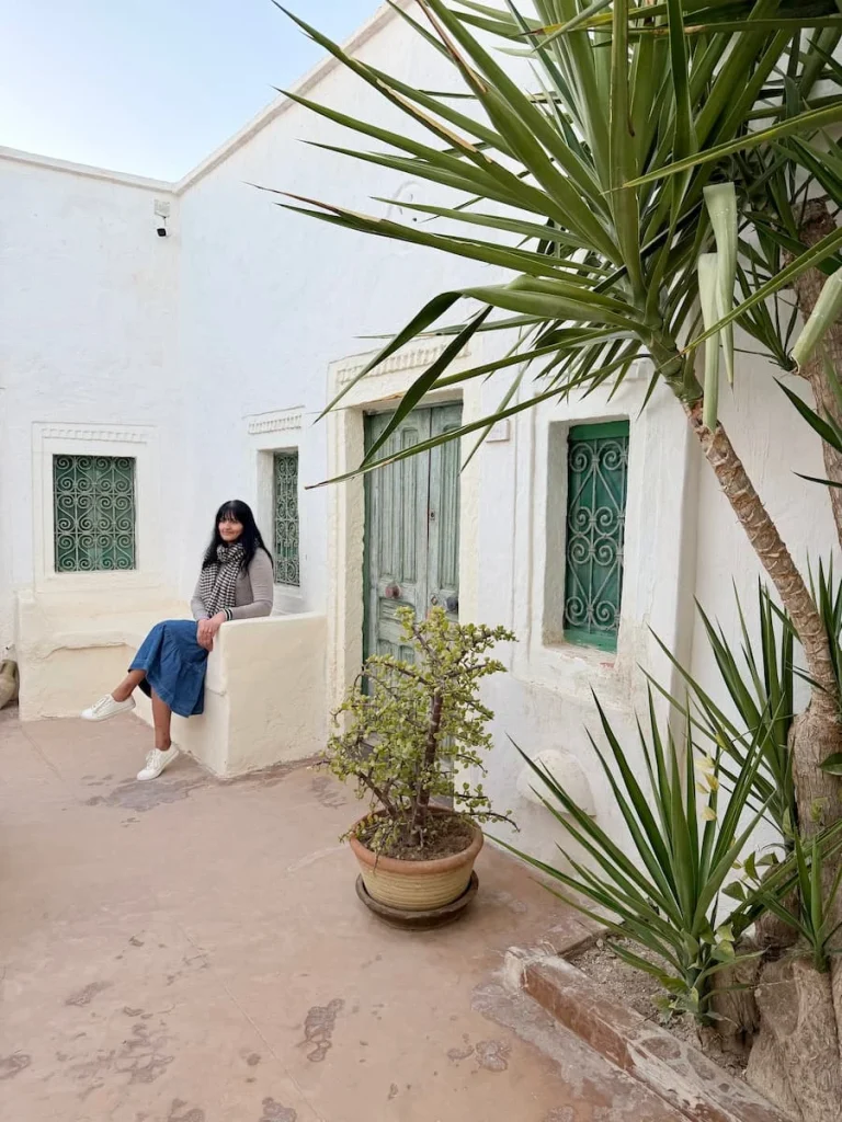Exterior of Dar Dhiafa room in Djerba, Tunisia Bejal is sitting on a white eat in the courtyard of the hotel with sage green doors and small palm plants framing the image