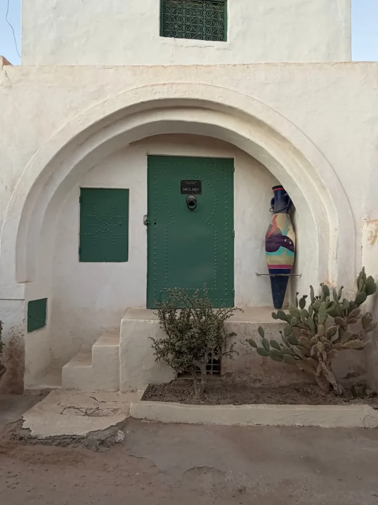 Green front door to Dar Dhiafa with a window beside it, a sign and cacti and potted plants outside, Djerba