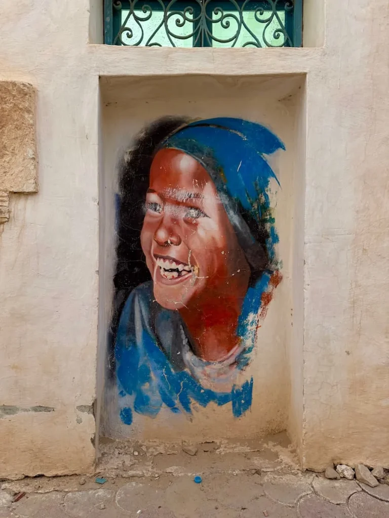 Girl street art in Djerbahood. The girl is a local wearing a blue hat and top , she has black hair. The artwork is painted on a cream wall in Djerba