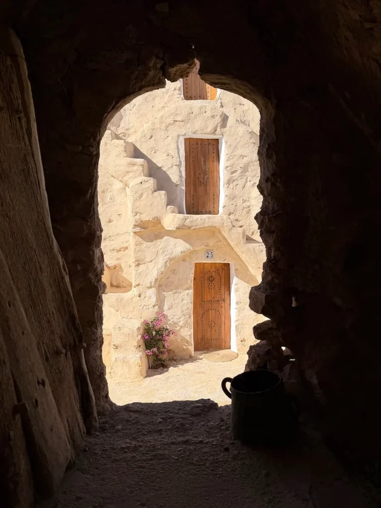 Ksar Hedada hotel doors through archway made of bricks and sand. The image takes a peek at a floral plant and two level of rooms