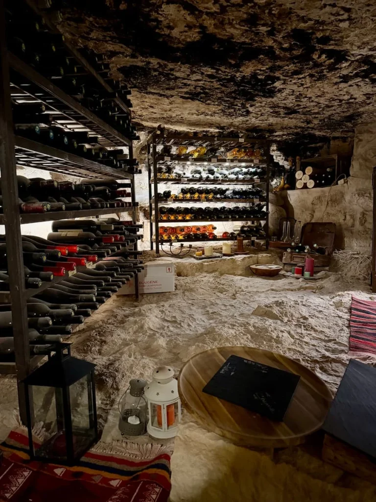 L'Auberge de Tamezret wine cellar. There are various bottles on wine stocked up on a wine rack in teh cave like structure with rugs and other utensils in the soace.