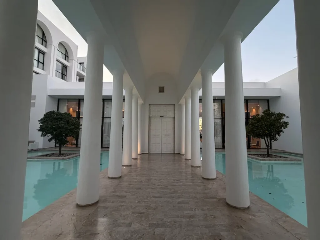 The white pillars of the La Badira entrance. There is a middle alleyway with floor to ceiling pillars and at either side are ponds with trees on a small square island base.