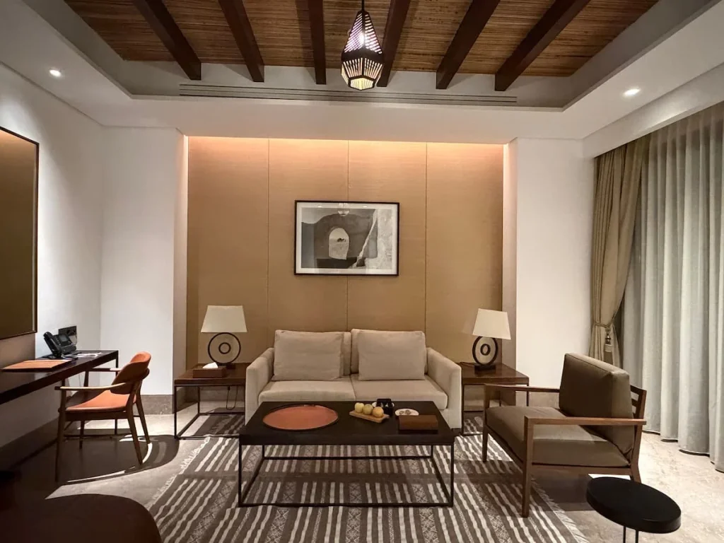 Lounge area in the villa at The Mora, Tozeur. There is a beige sofa in the middle of the room with a black coffee table, lamps to the side and a console table to the left with a red cusioned chair. The coffee table has a traditional Tunisian rug under it