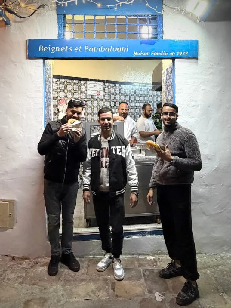 3 men at the Bamboulini stall, Sidi Bou Said, Tunisia. They are standing outside the hole in the wall , wearing casual clothes tucking into the traditional doughnuts