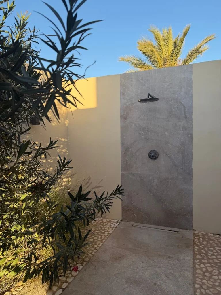 Outdoor shower at The Mora, Tozeur. The shower is on against a light cream stone background with foliage and plants to the left hand side