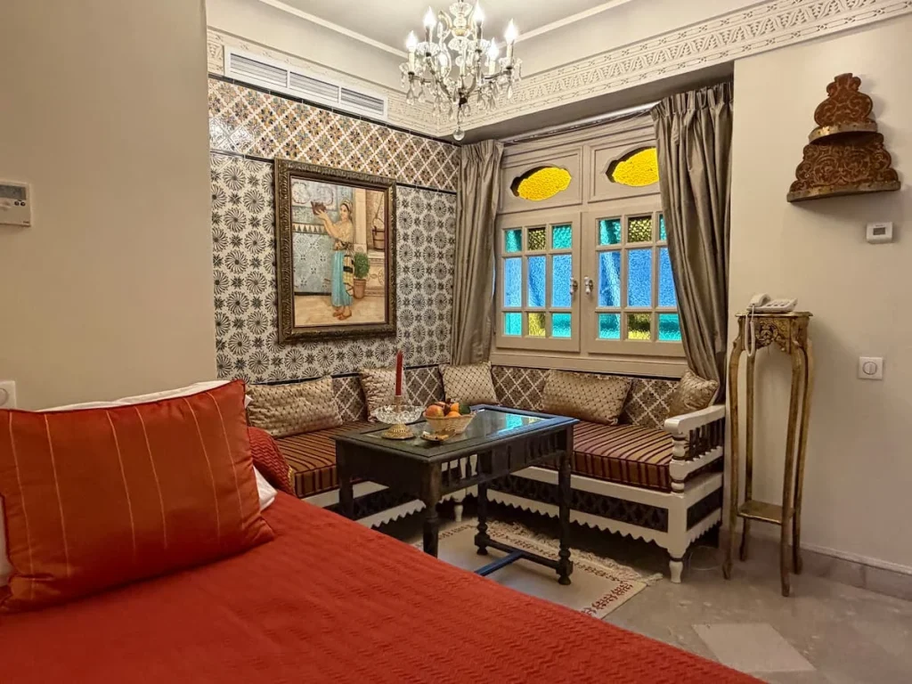 Seating area and extra day bed in suite at Dar Alouini, Kairouan, Tunisia. There is a table and padded inlaid seating area with fruit and Tunisian date suites and a red covered day bed to the left.