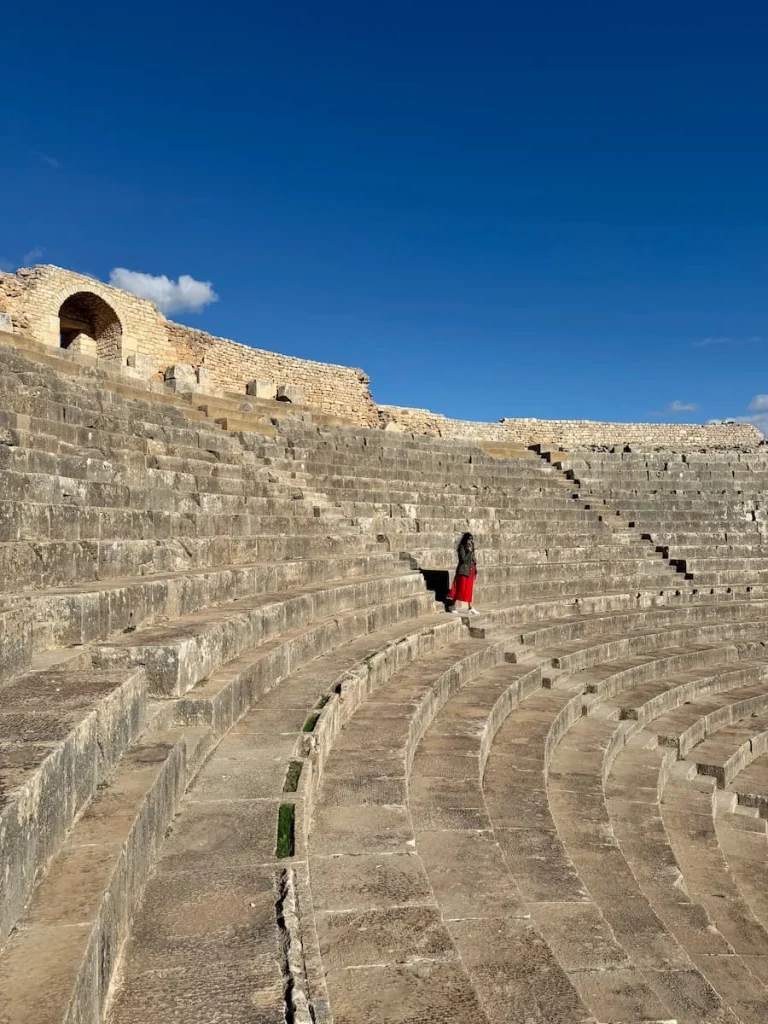 Side view of Dougga amphitheatre, Tunisia. Bejal is walking dow the tiered seating wearing a red skirt and black top
