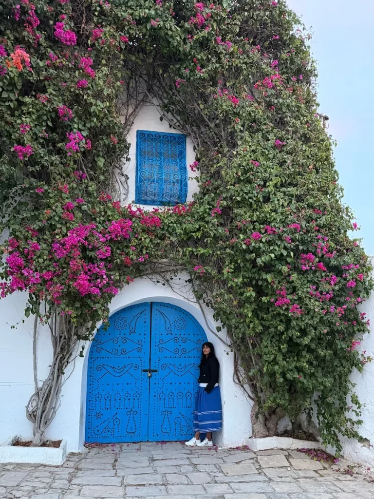 Sidi Bou Said blue door with foliage and bougainvillaea, Tunisia. Bejal is wearing a white top and blue skirt standing against the door