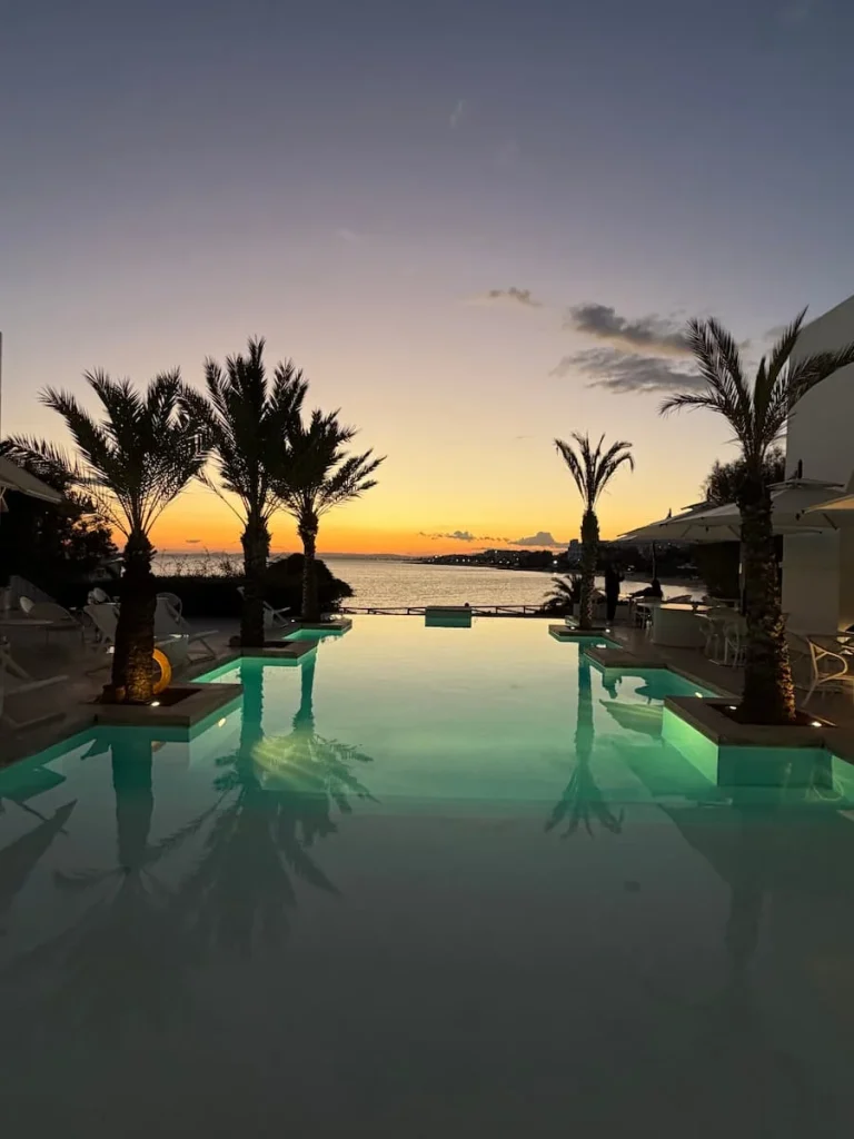 Sunset at La Badira over the infinity pool and palm trees swaying in the background. The sky is an orange and yellow colour. La Badira is located in Hammamet