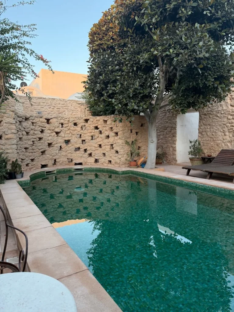 Swimming Pool at Dar Dhiafa with green water and a beige wall at the back with geometric sqaure carved out of it