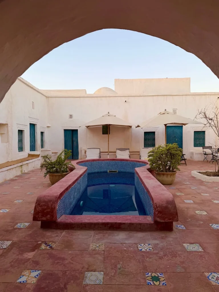 Swimming pool at Dar Dhiafa with sun loungers, a tan tiled floor and rooms coming off it. The whole photo is framed by white stone work in Djerba