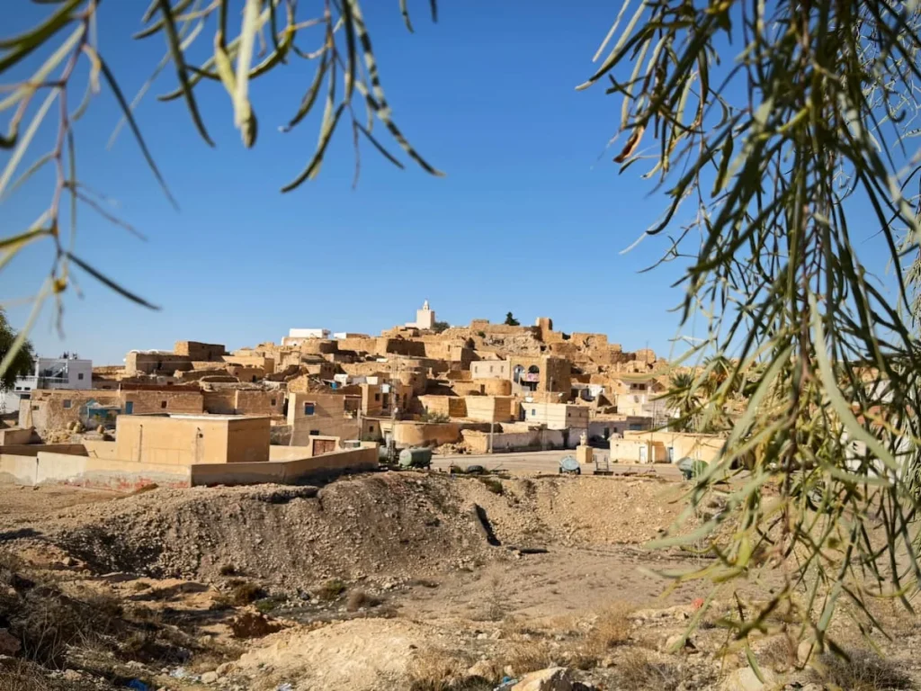 A landscape photo of the small Berber town of Tamezret, Matmata, Tunisia. The mountain town is all colours of san, white and brown with the image being framed by green tree foliage