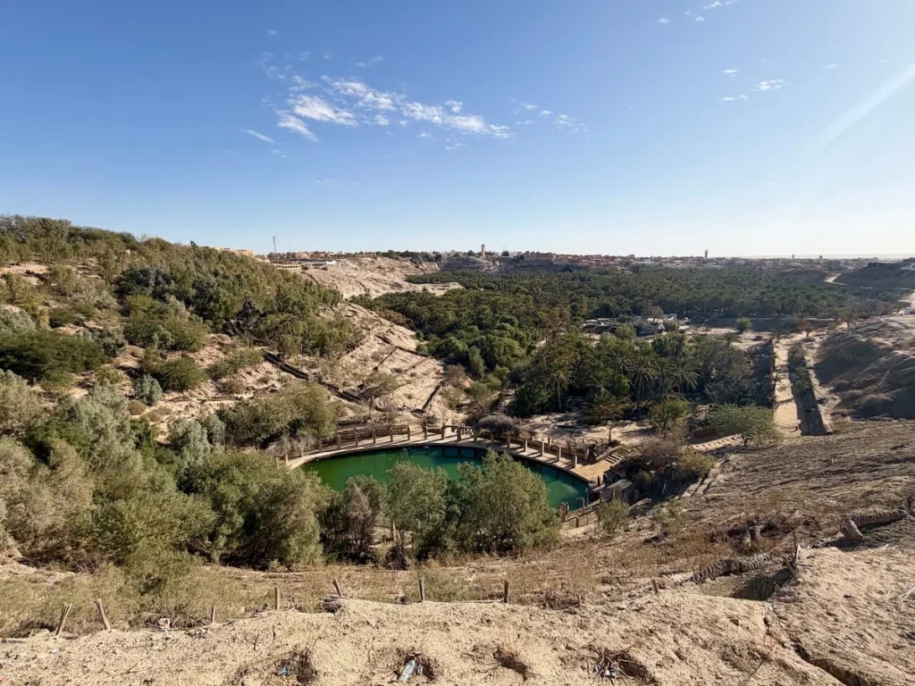 Tozeur Oases in Tunisia. The central lake is of a green blue colour and is surrounded by palm trees and a rocky terrain.