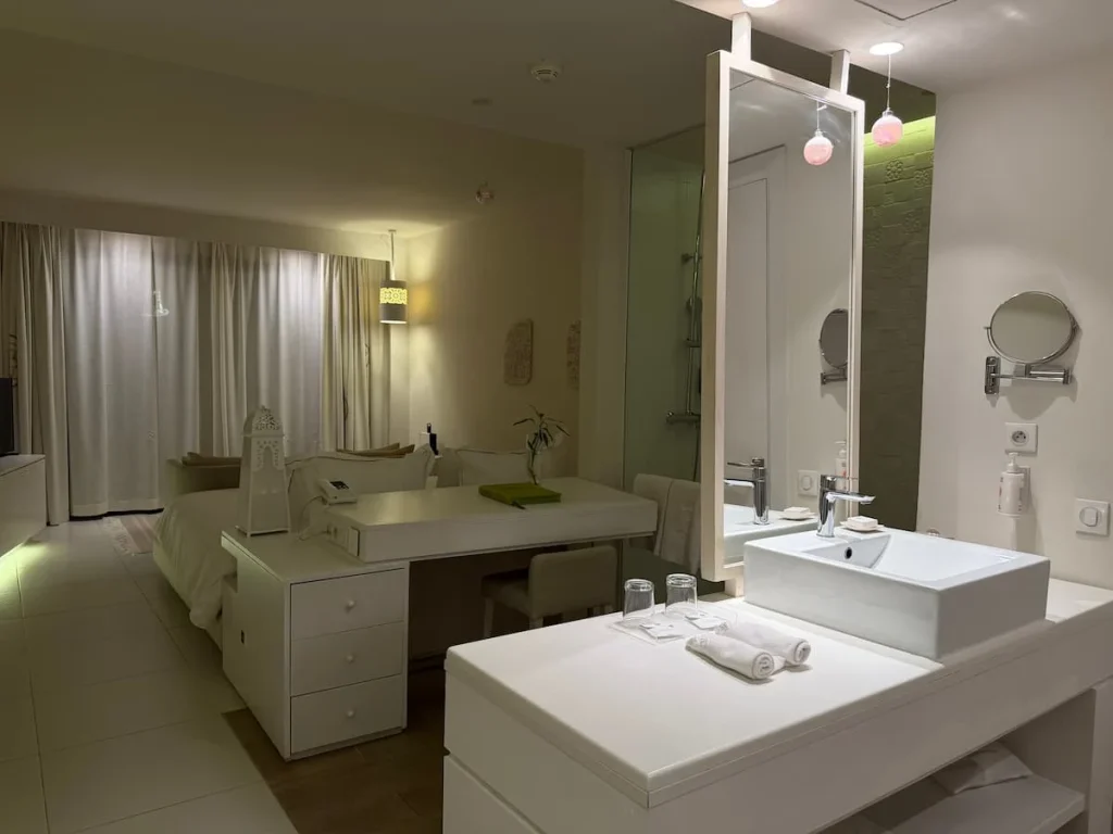 Interiors of suite at La Badira with the vanity unit, bed and sofa and shower. The image is taken in the evening - the room is white with low atmospheric lighting
