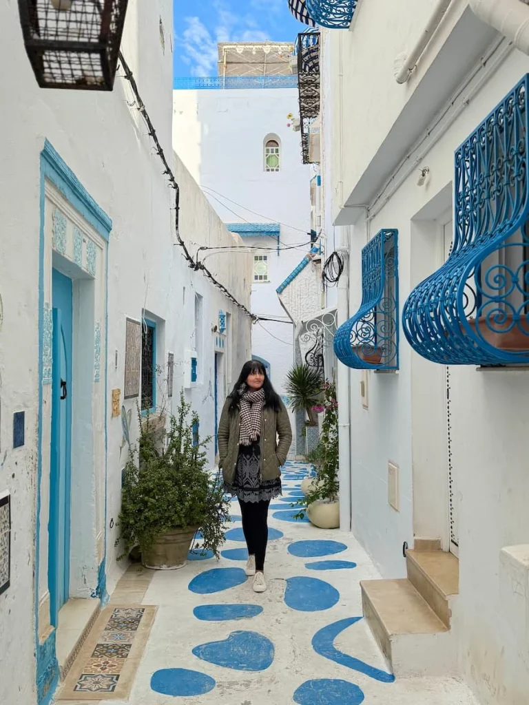 Bejal in Hammamet Medina walking along a white washed alleyway with blue patches on the floor adn blue grid windows adn doors
