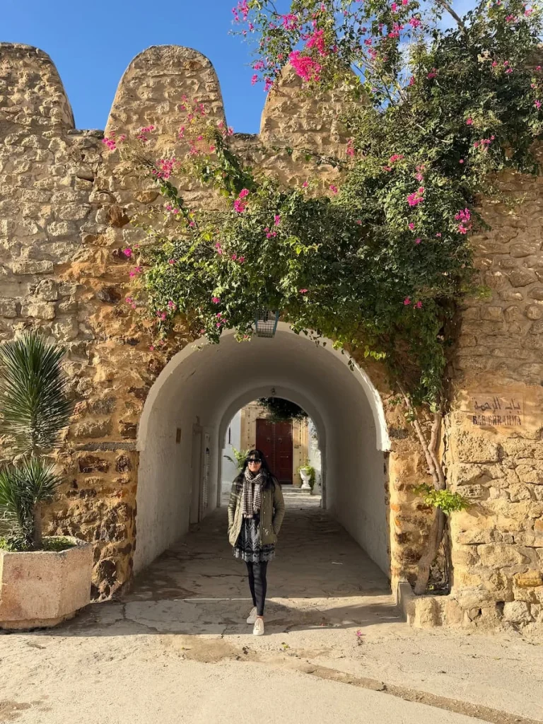 Bejal walking through Archway into Hammamet Medina which is made from stone and surrounded by plants and small pink flower. Bejal is wearing black leggings a black cut-work dress adn a khaki padded jacket